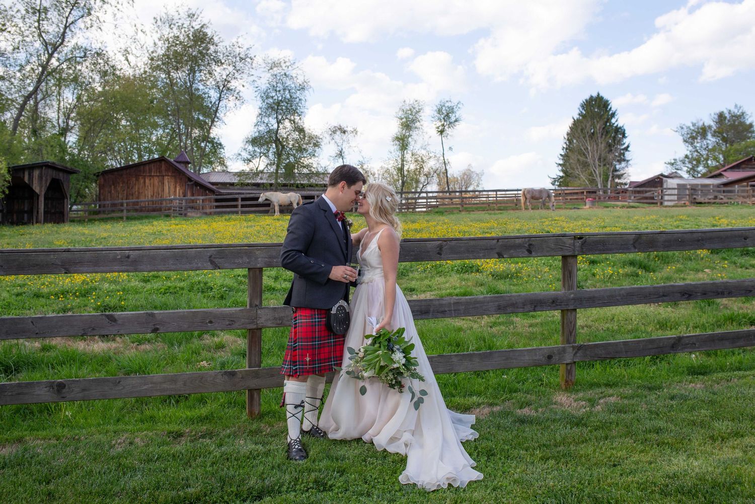 Bride and groom in Scottish kilt share a romantic moment by a rustic wooden fence at a countryside wedding venue.