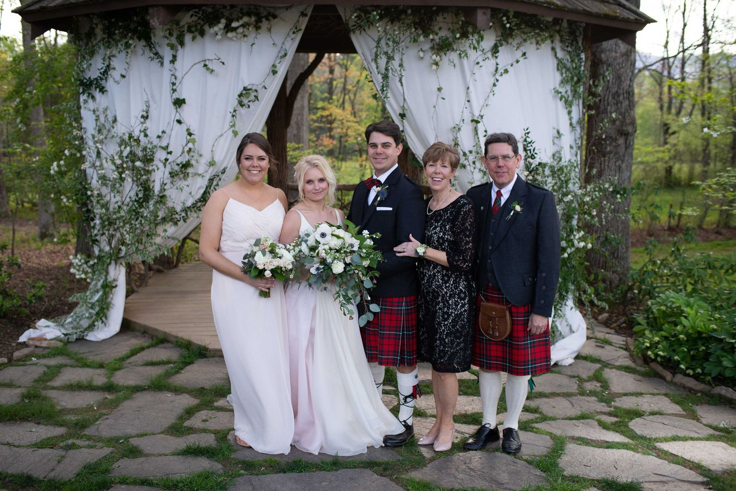 Wedding party poses under floral gazebo, featuring kilts and bridal gown in an outdoor garden ceremony.