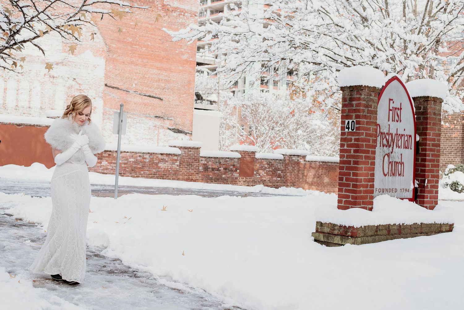 Bride in white gown stands in snowy winter scene beside First Presbyterian Church brick sign.