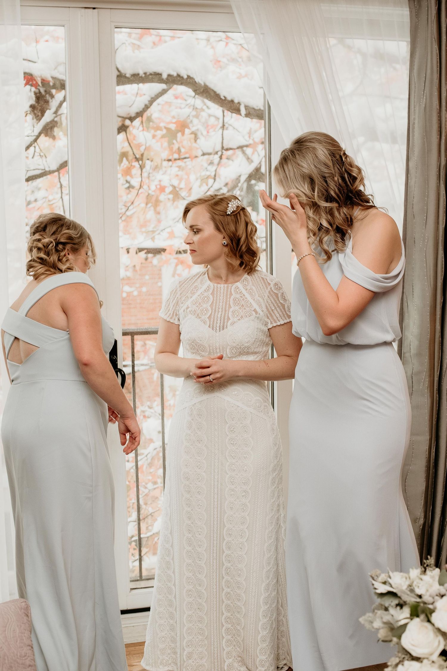 Bride in lace gown stands between two bridesmaids in silver dresses near a window during wedding preparations.