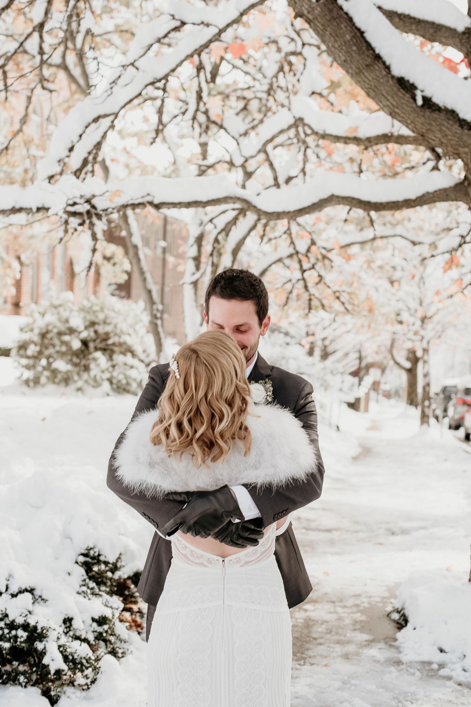 Bride and groom embrace in a snowy winter wonderland, bride wearing a white fur shawl over her wedding gown.