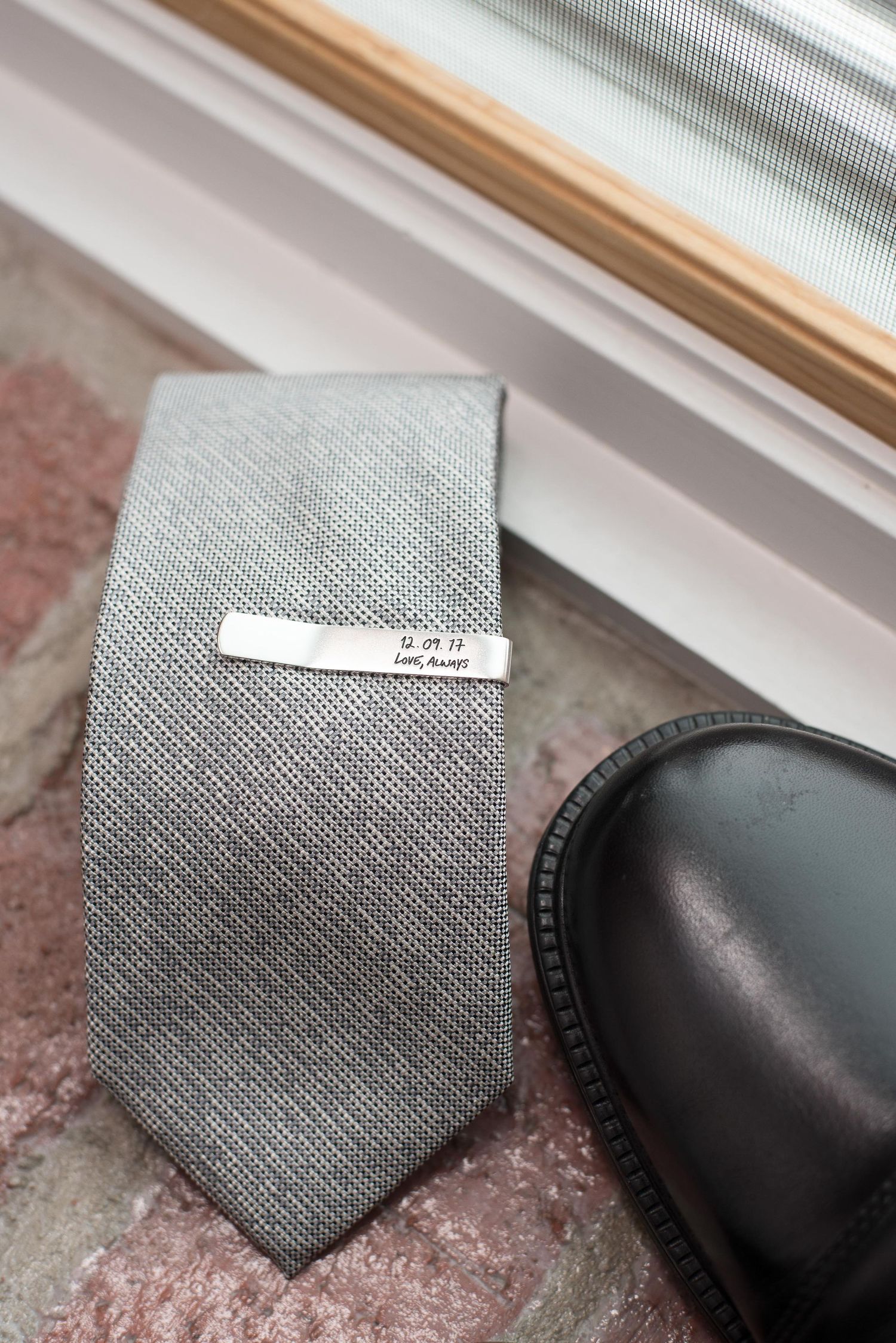 A gray silk tie with a personalized silver tie bar rests next to a black leather dress shoe on a marble surface.