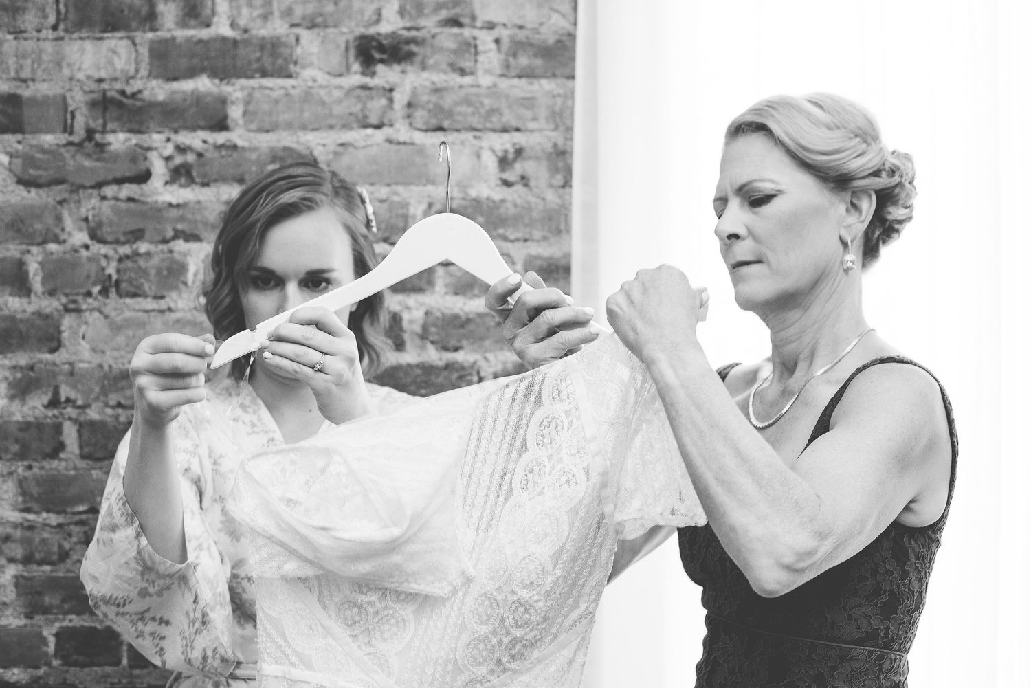 Black and white photo of bride and woman preparing wedding dress with lace details near brick wall.