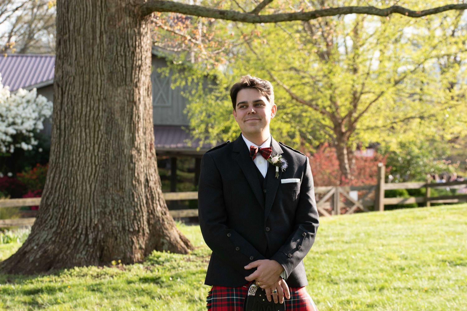 Man in black blazer and red tartan kilt stands smiling outdoors near large tree at a wedding venue.