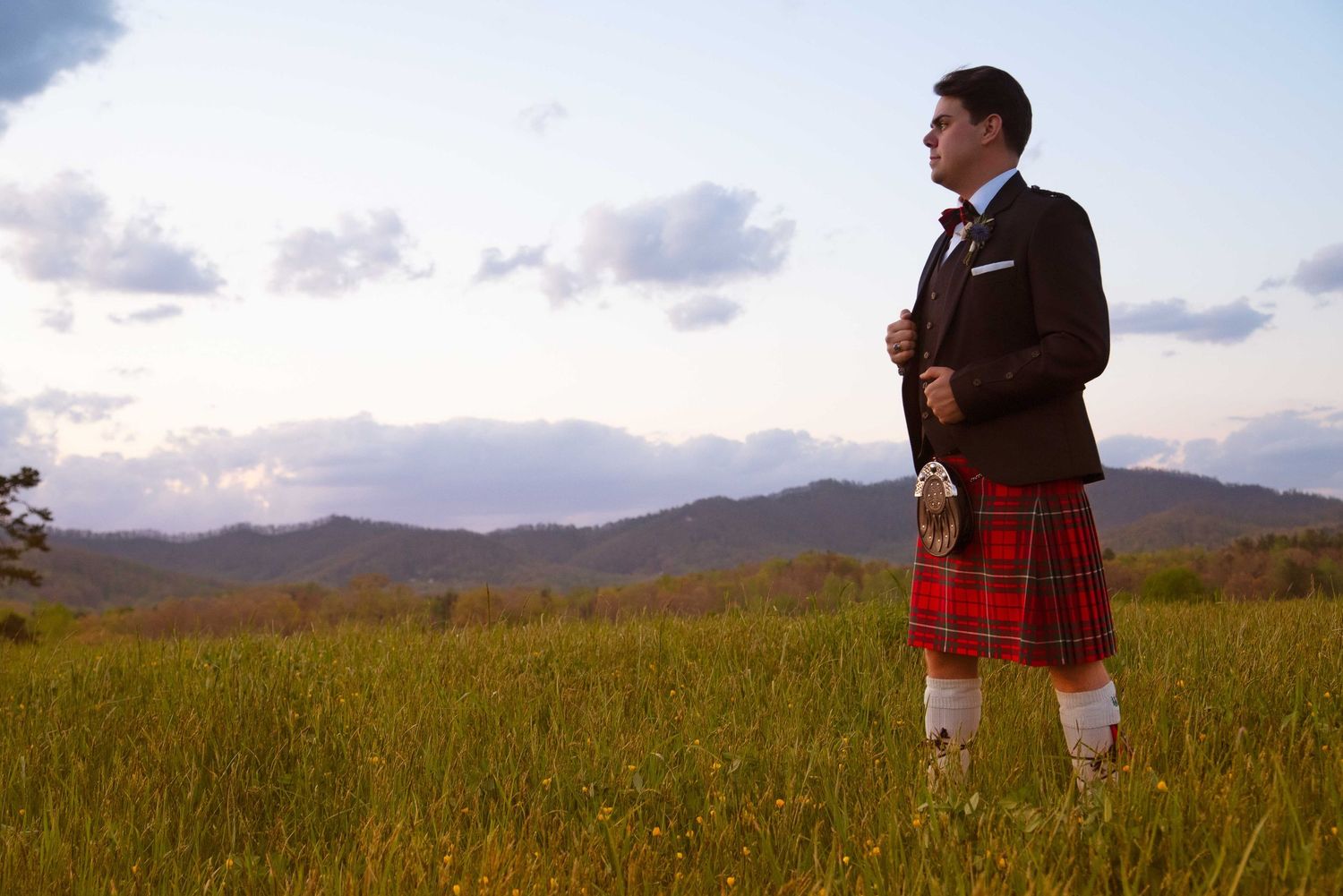 Man in traditional Scottish kilt and blazer standing in green field with mountain backdrop at dusk.