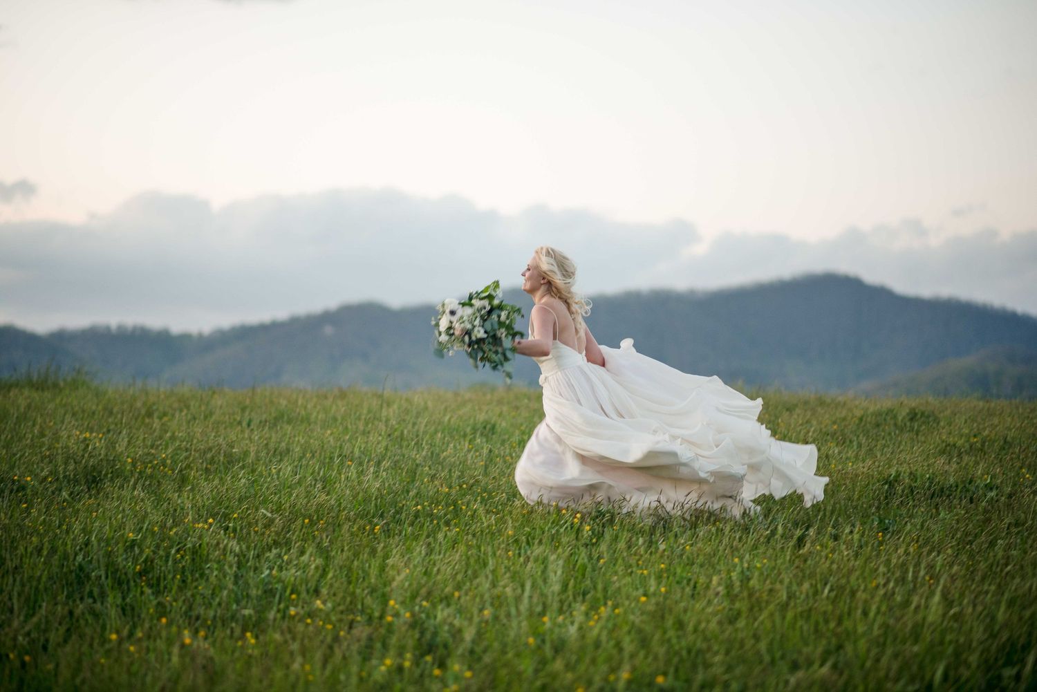 Bride in flowing white gown runs through green mountain meadow holding bouquet on wedding day.