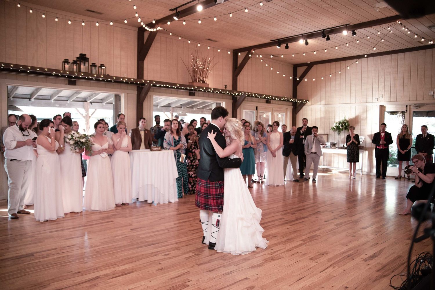 Bride and groom share first dance in rustic barn wedding venue surrounded by bridesmaids and guests with string lights.