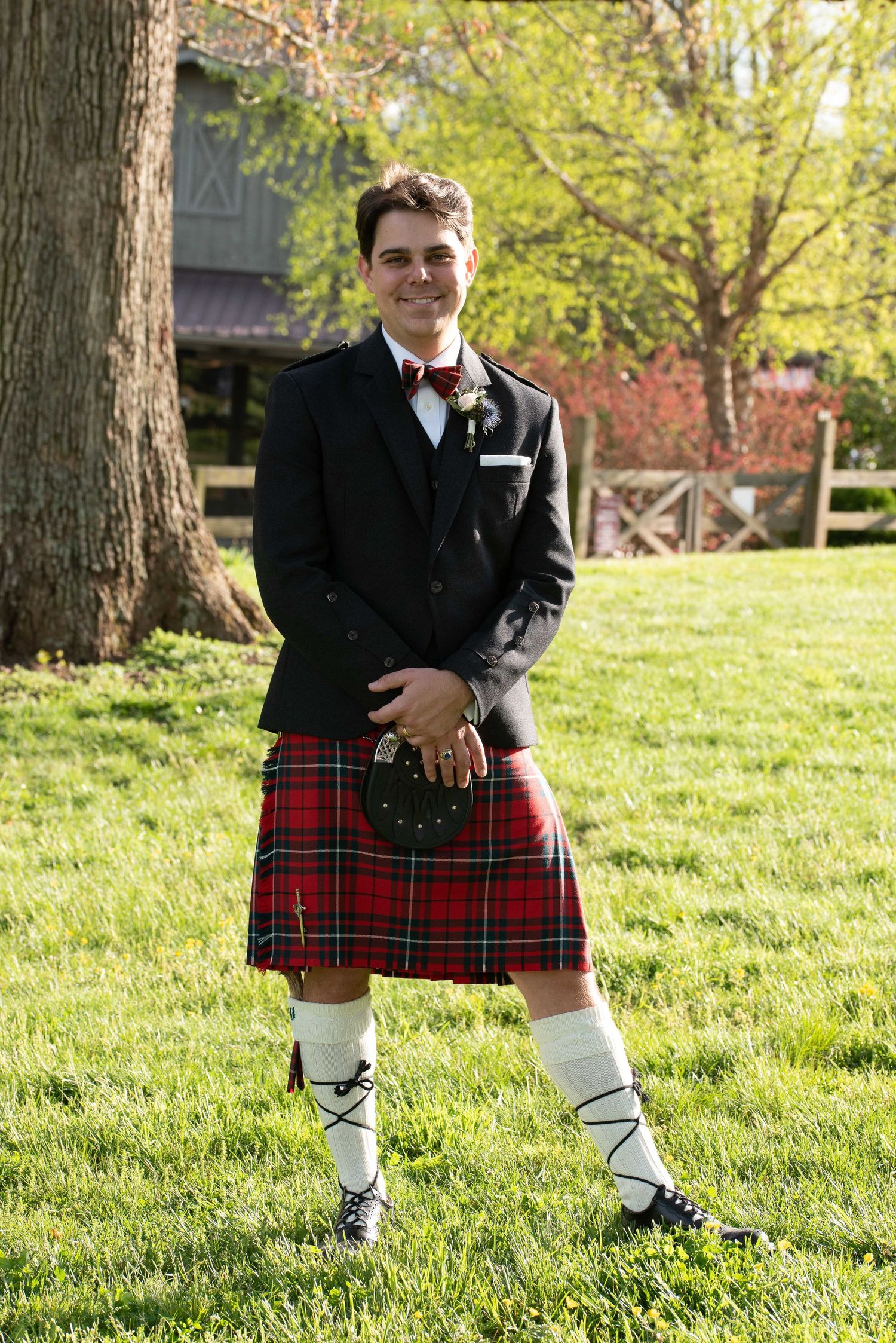 Man in traditional Scottish kilt outfit with black jacket, red tartan, sporran, and white knee socks standing on green grass.