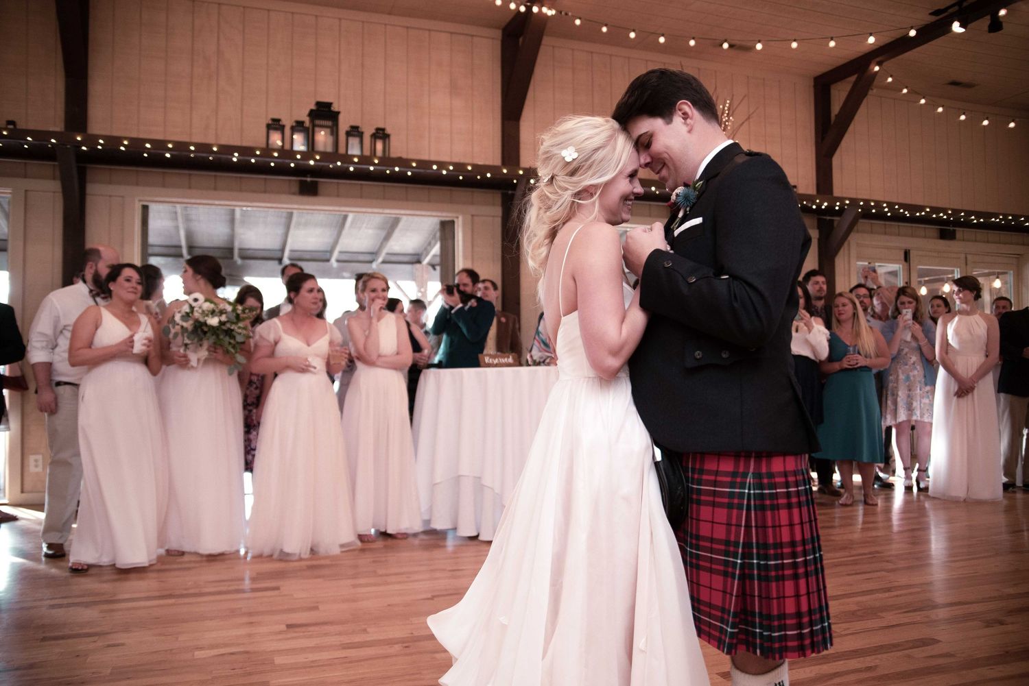 Bride and groom share first dance at rustic barn wedding, groom wearing traditional Scottish tartan kilt, bridesmaids in blush gowns.