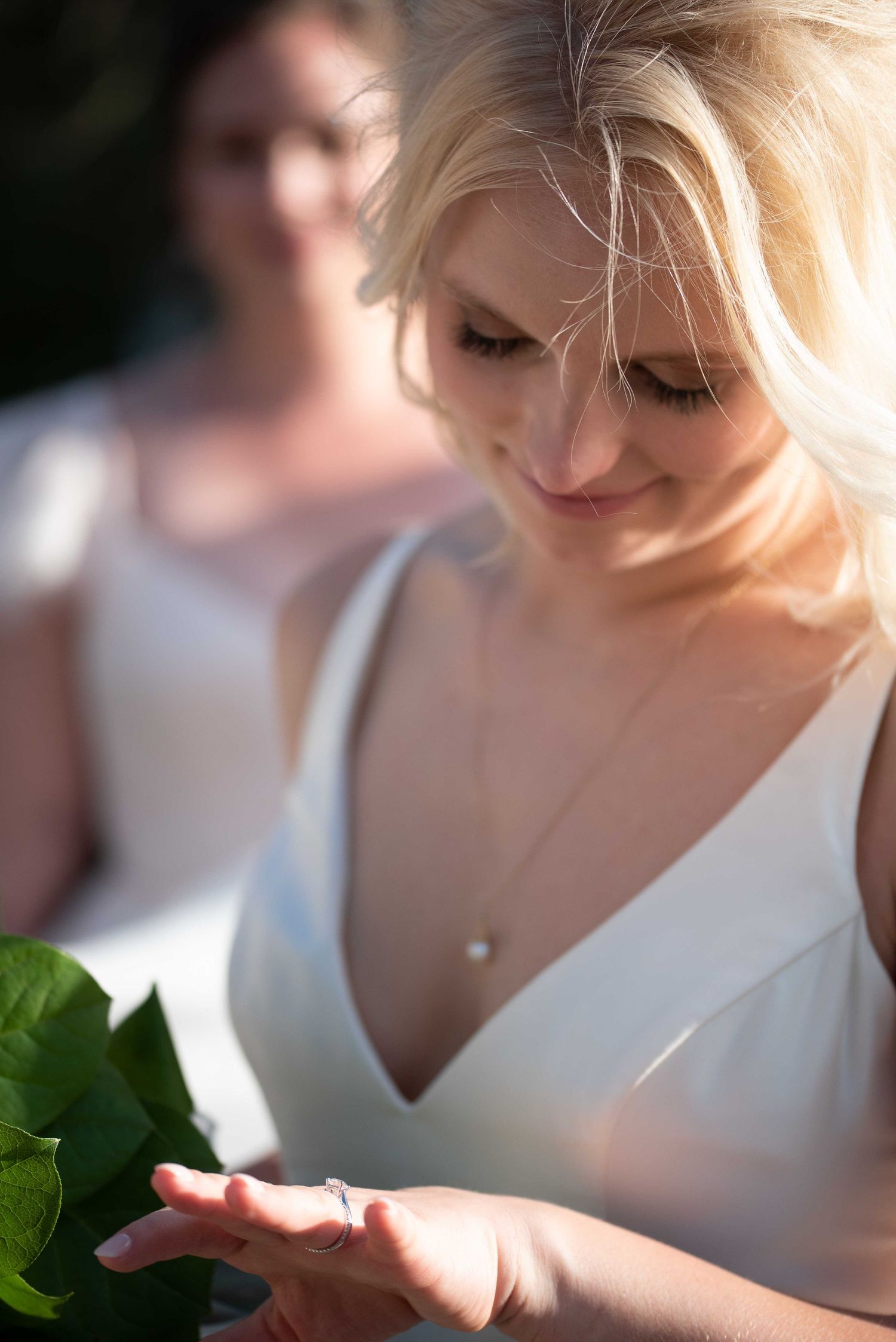 Smiling blonde bride in white V-neck dress looking down at her wedding ring outdoors.