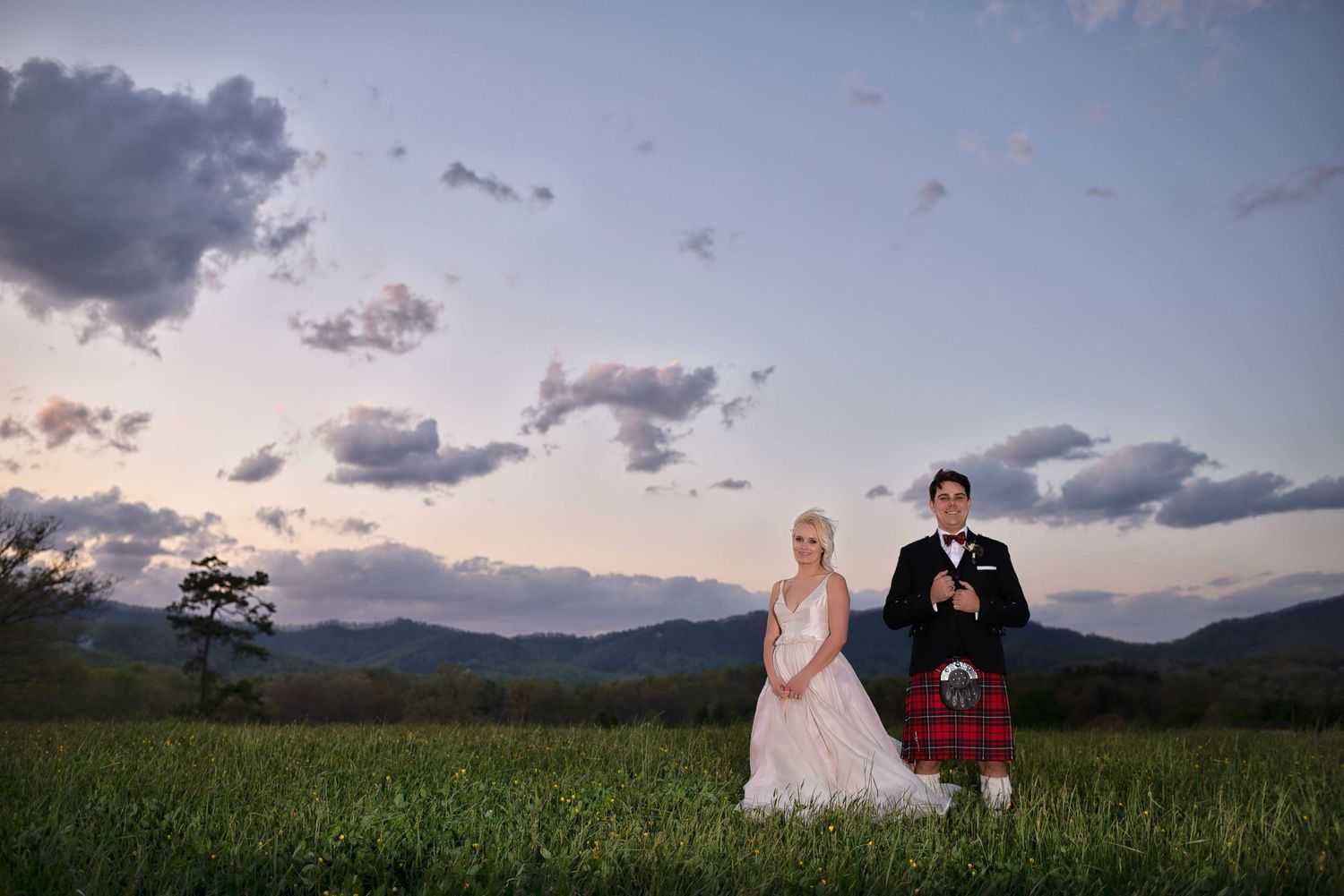 Bride in white gown and groom in red tartan kilt stand in green field at sunset with mountain backdrop.