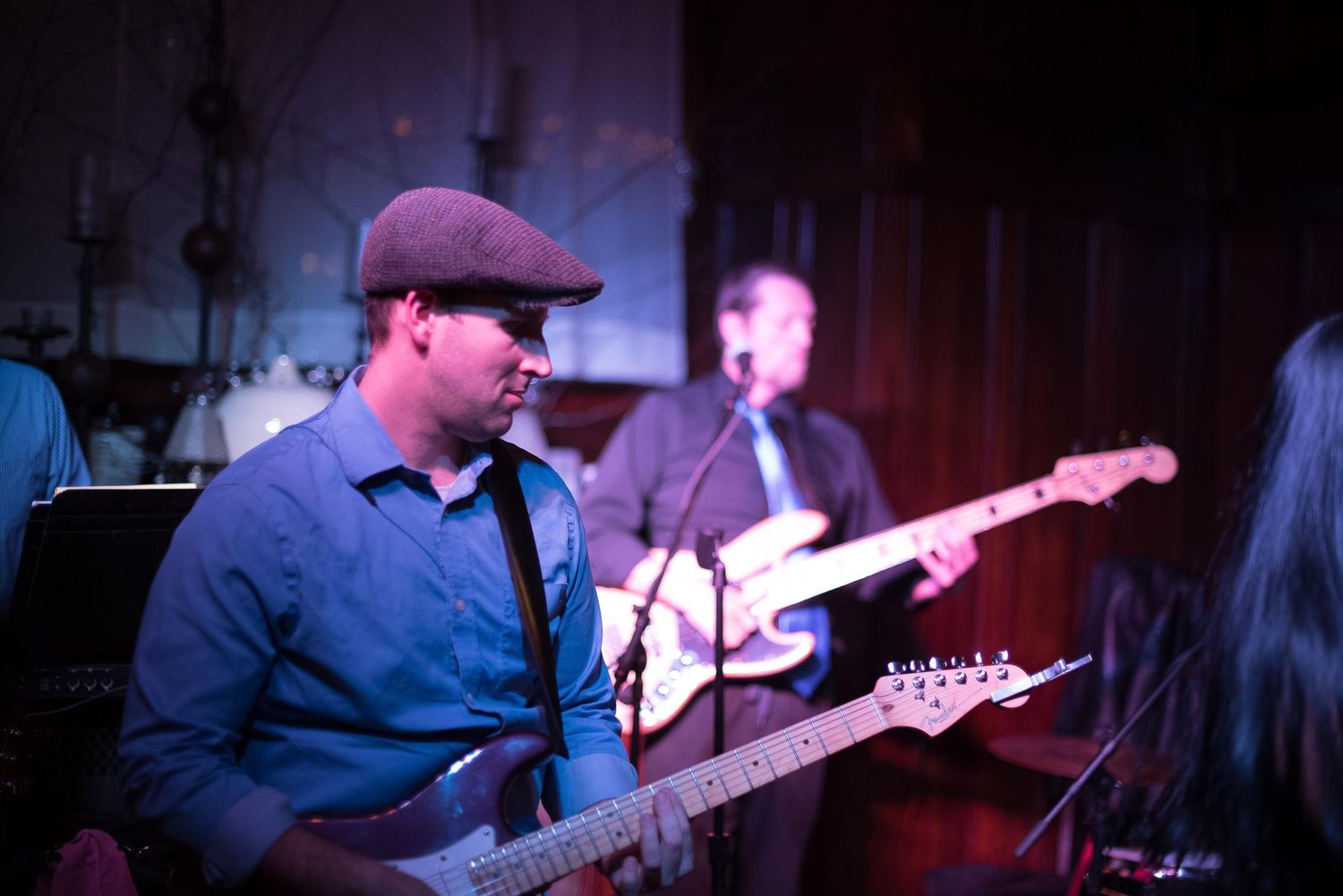 Live band performance with guitarist in blue shirt and cap playing electric guitar on dimly lit stage.