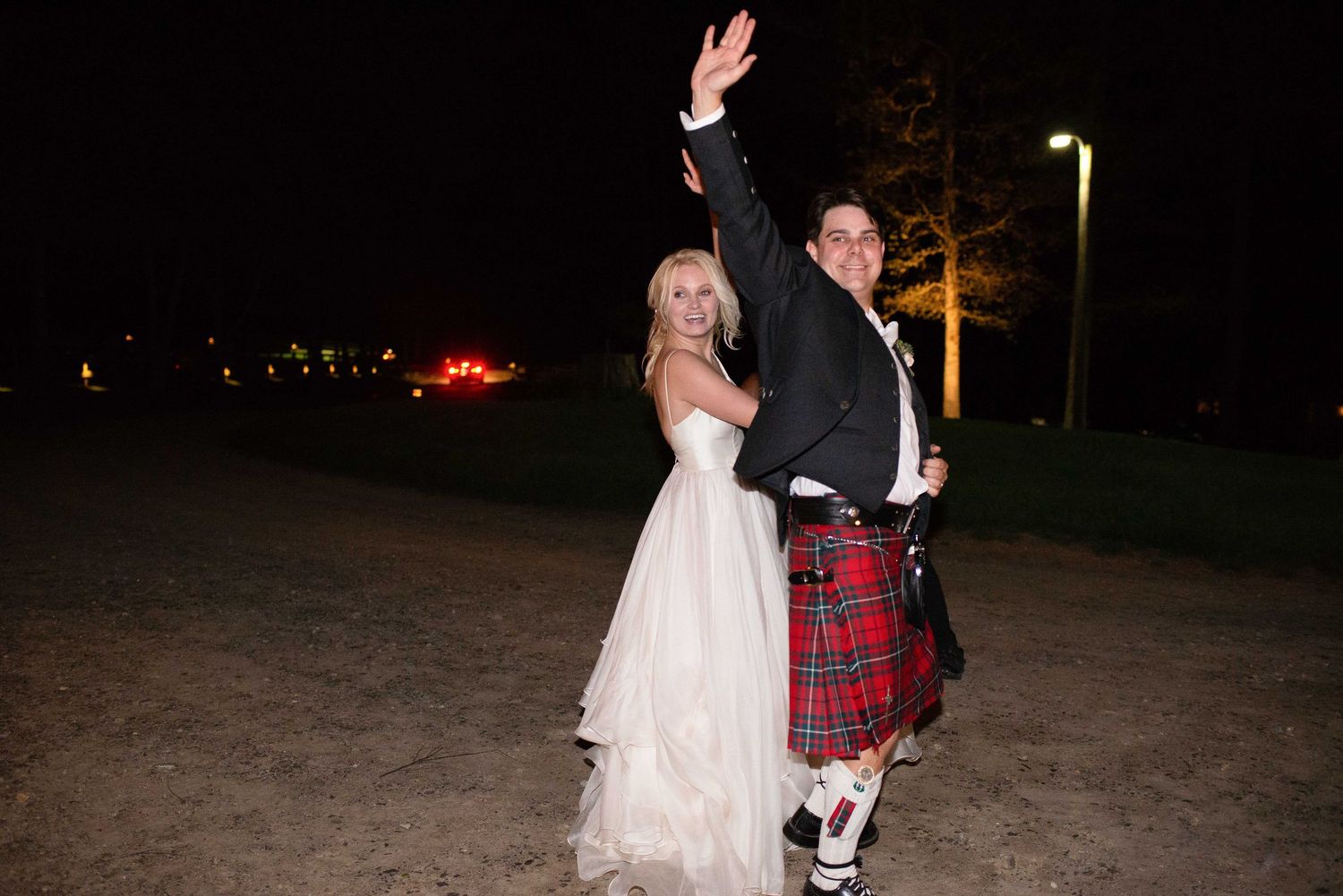 Bride in white gown and groom in red tartan kilt waving at night outdoor wedding celebration.
