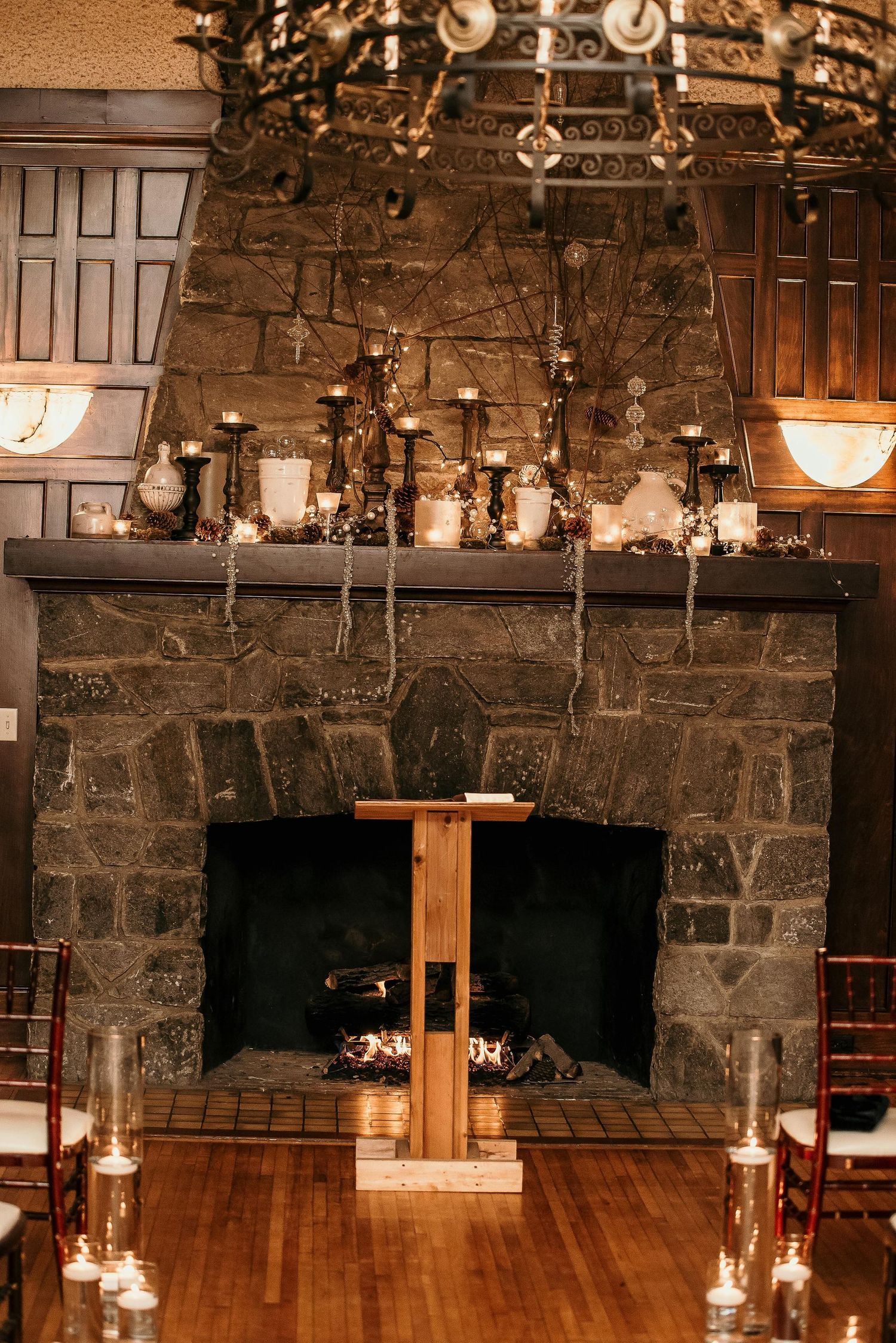 Rustic stone fireplace ceremony backdrop with glowing candles, wooden podium, and ornate iron chandelier above.