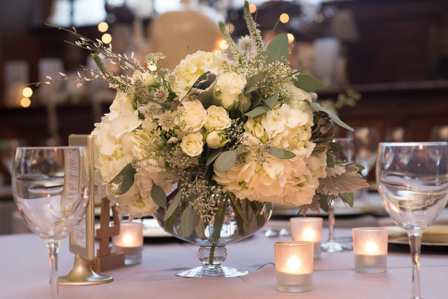 Elegant wedding centerpiece with white hydrangeas and roses in a silver bowl, surrounded by glowing votive candles on a white tablecloth.