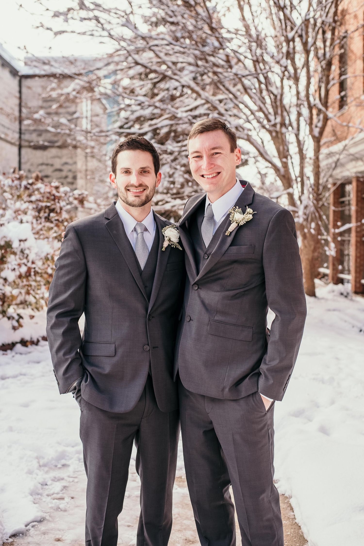 Two grooms in matching gray suits with boutonnieres posing outdoors in a snowy winter wedding setting.
