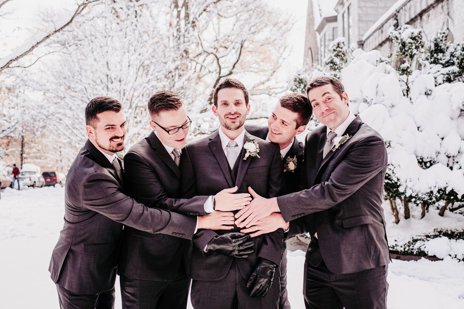 Groom and groomsmen in dark suits laugh together outdoors in snowy winter wedding setting.