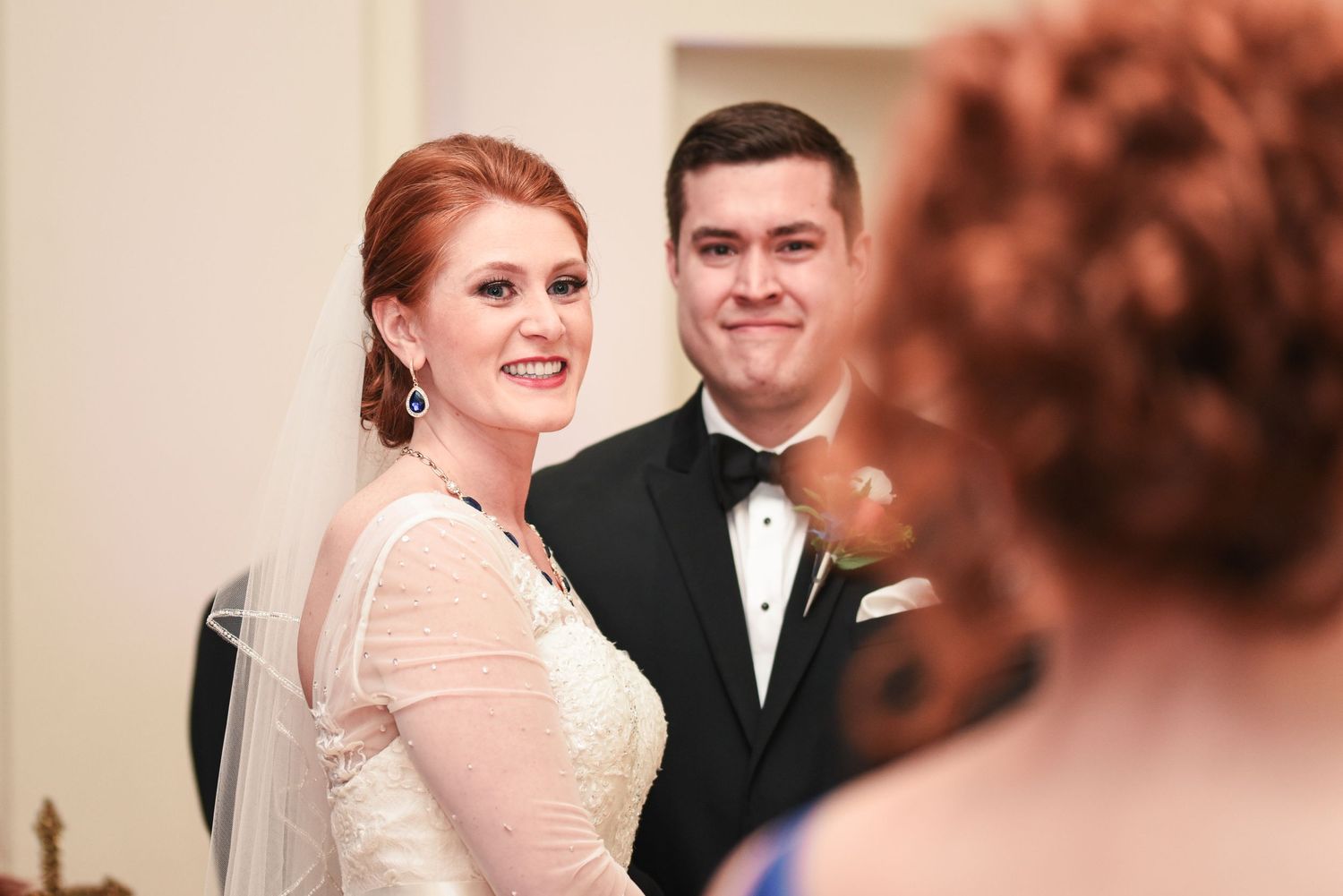 Smiling red-haired bride in lace gown and groom in tuxedo sharing a joyful moment during wedding ceremony.