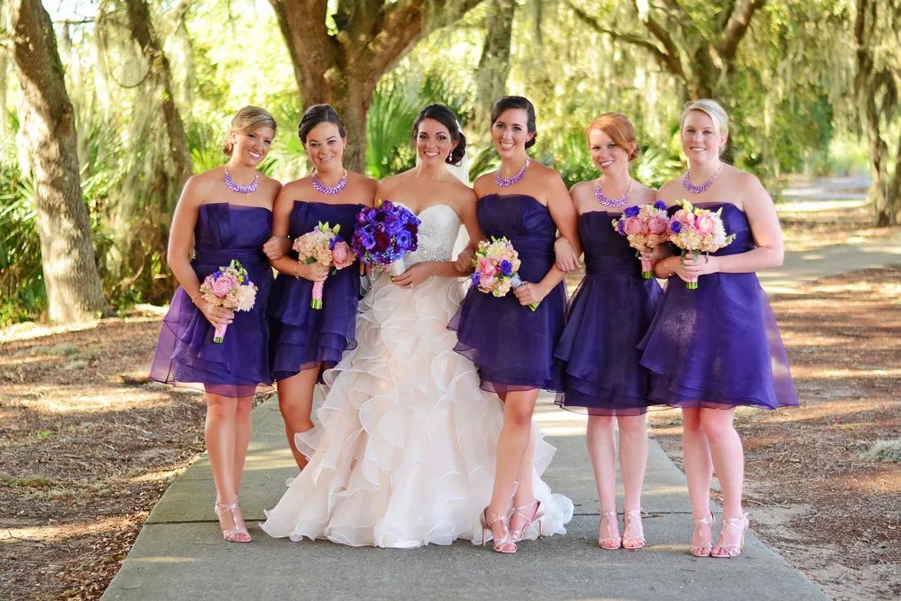 Bride in ruffled white gown poses with five bridesmaids in purple strapless dresses holding colorful bouquets outdoors.