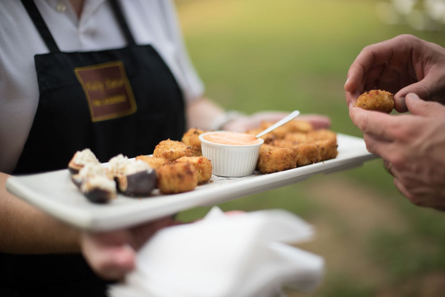 Caterer serves golden fried appetizers with dipping sauce on a white tray at an outdoor wedding reception.