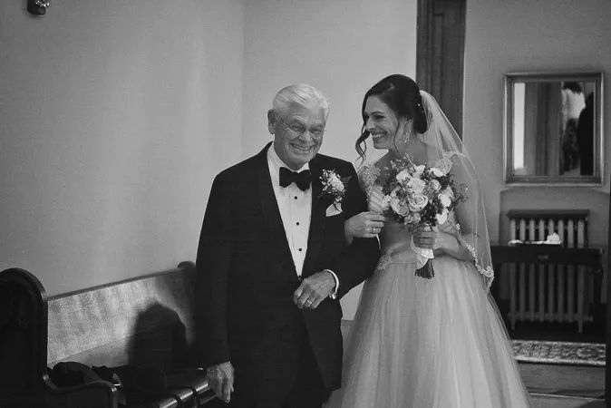 Black and white photo of smiling bride walking with elderly father in tuxedo before wedding ceremony.