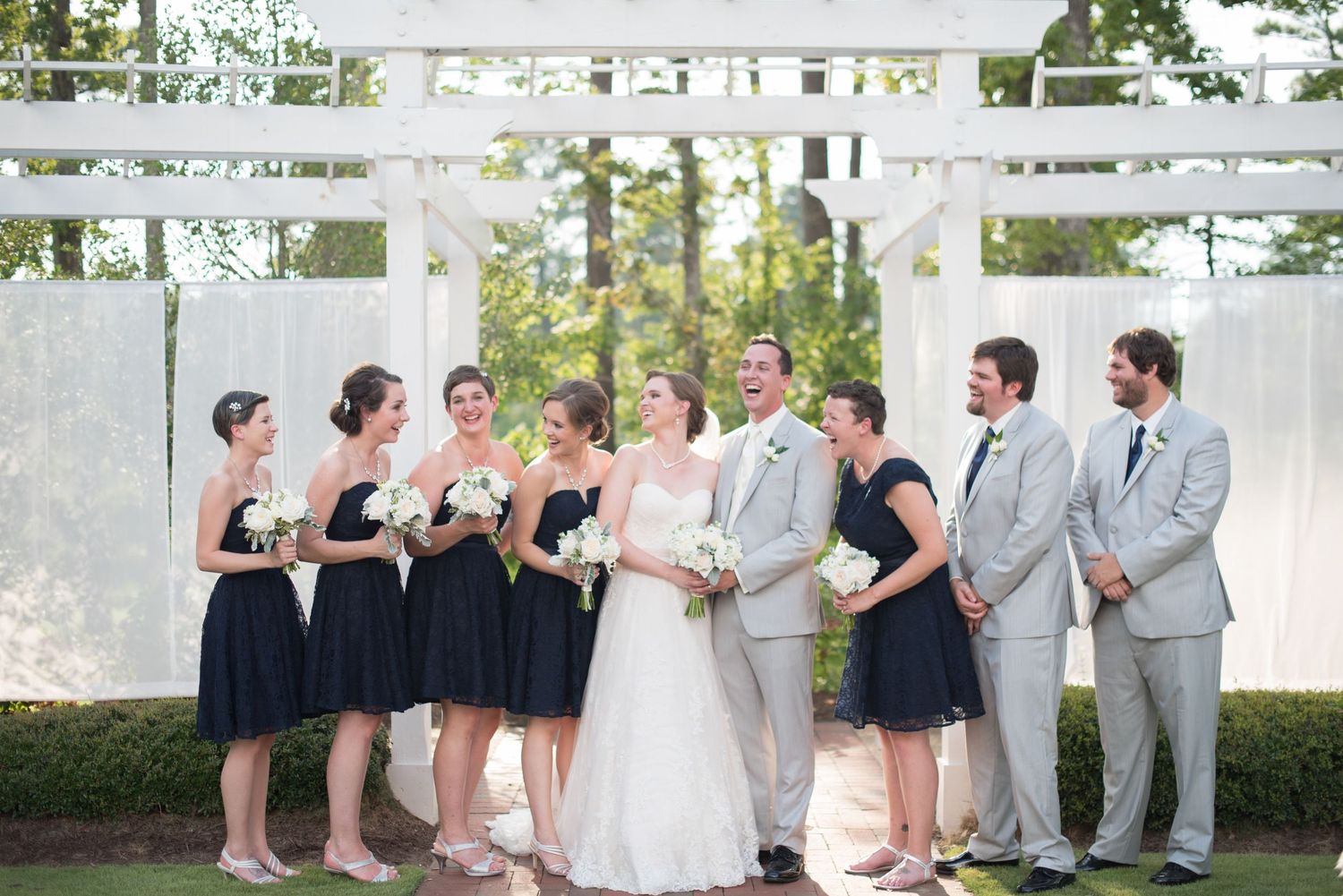 Wedding party in navy and grey attire smiles together outdoors near a white pergola backdrop.