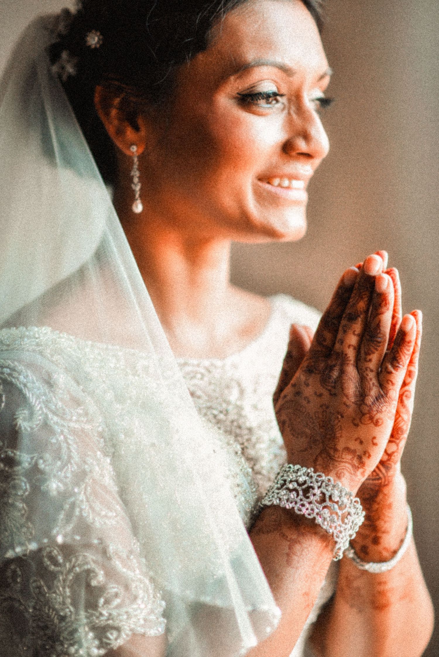 South Asian bride in white lace gown with henna hands in prayer pose, wearing veil and pearl earrings.