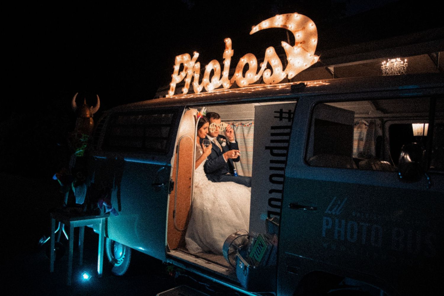 Bride and groom posing inside a vintage VW bus photo booth with glowing marquee lights spelling Photos at night.