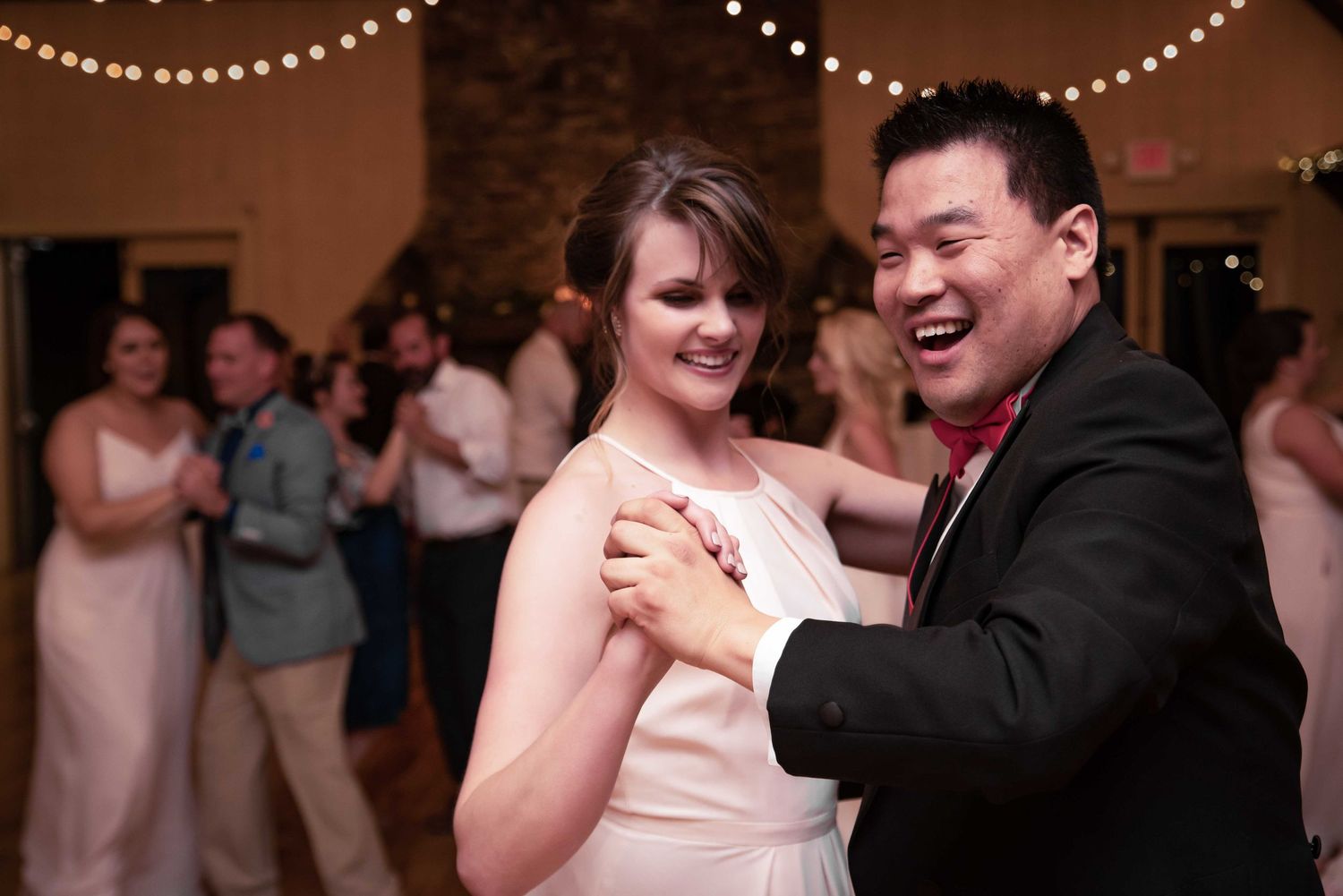 Couple sharing a joyful dance at a wedding reception with string lights and guests in the background.