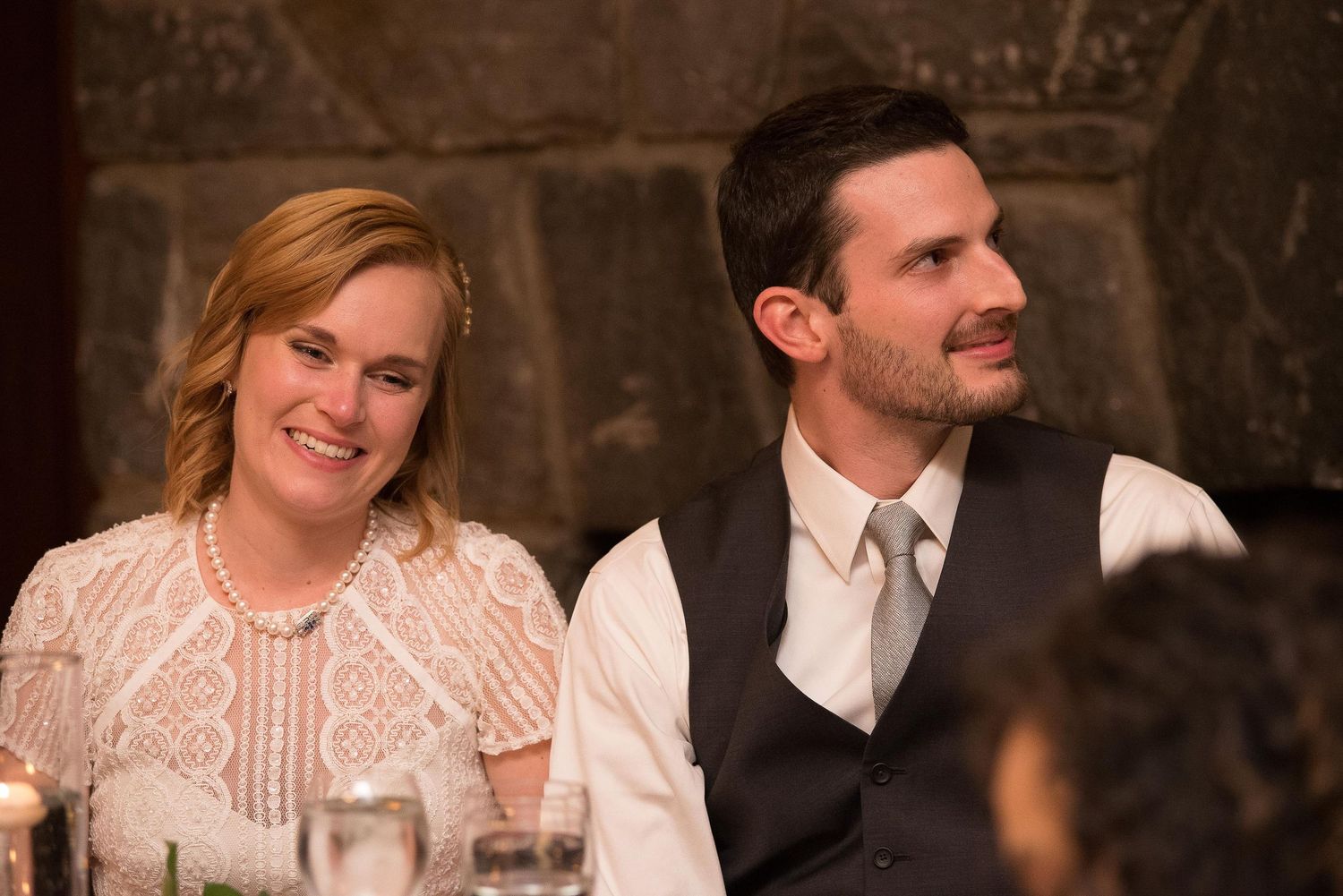 A smiling couple in formal attire seated at a wedding reception table, woman in white lace dress with pearls.