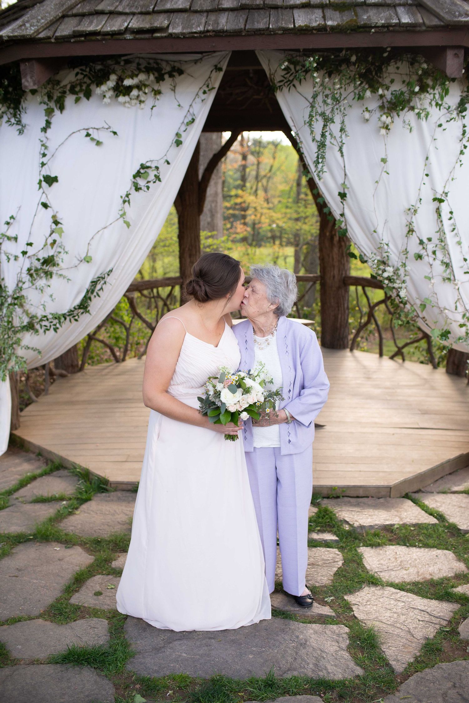 Bride in white gown holding bouquet shares a kiss with elderly woman in lavender suit under decorated gazebo.