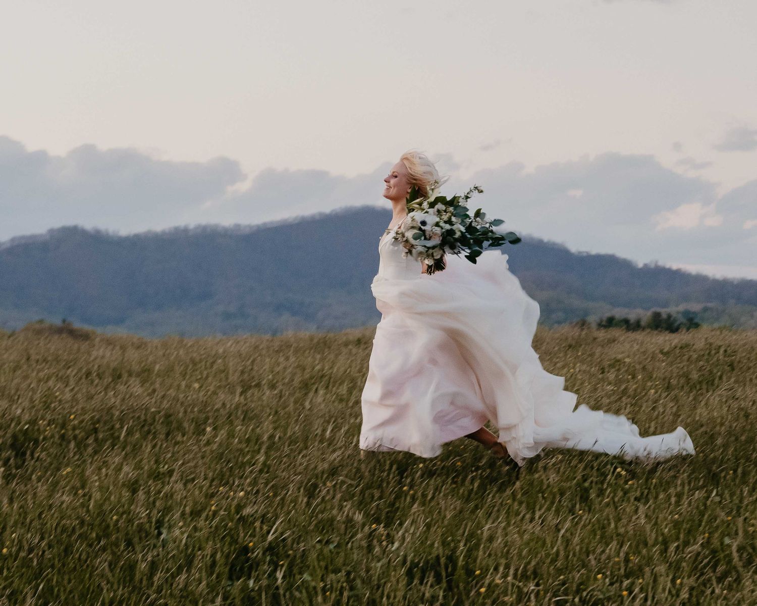 Bride in flowing white gown holding floral bouquet runs through green meadow with mountain backdrop.