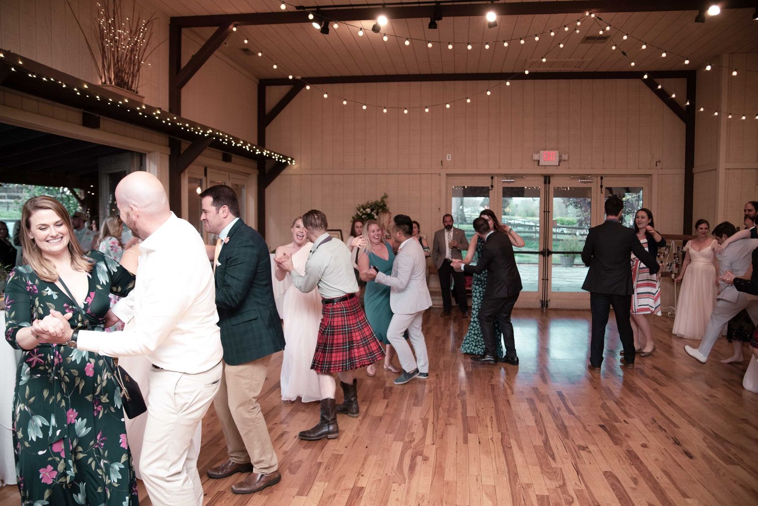 Guests dancing joyfully at a rustic barn wedding reception with string lights and hardwood floors.