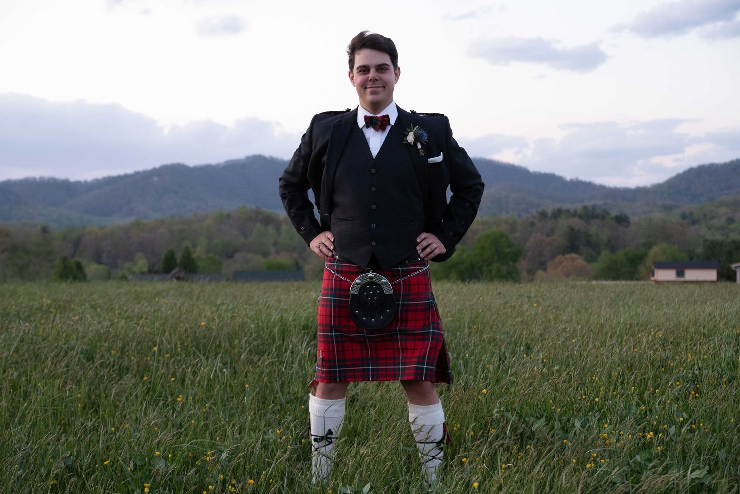Man in traditional Scottish attire with red tartan kilt and black jacket standing in green field with mountain backdrop.