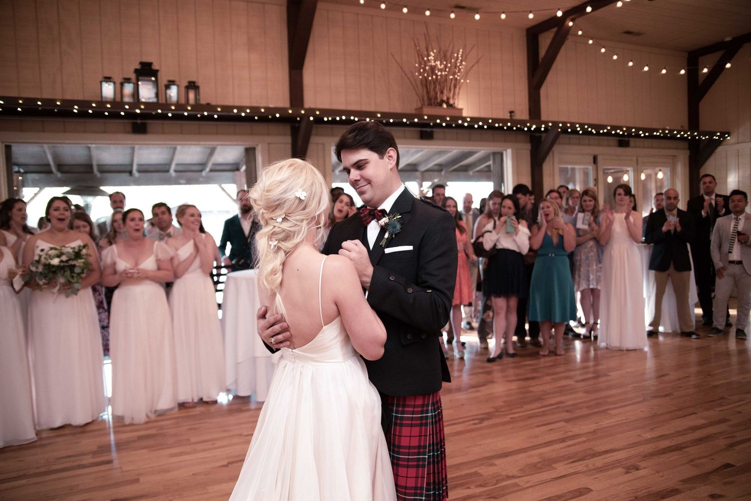 Bride and groom share their first dance in a rustic barn venue surrounded by bridesmaids in pink dresses.