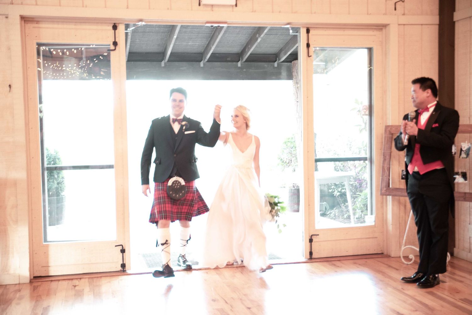 Bride and groom make grand entrance at wedding reception, groom wearing traditional Scottish kilt, greeted by emcee with microphone.