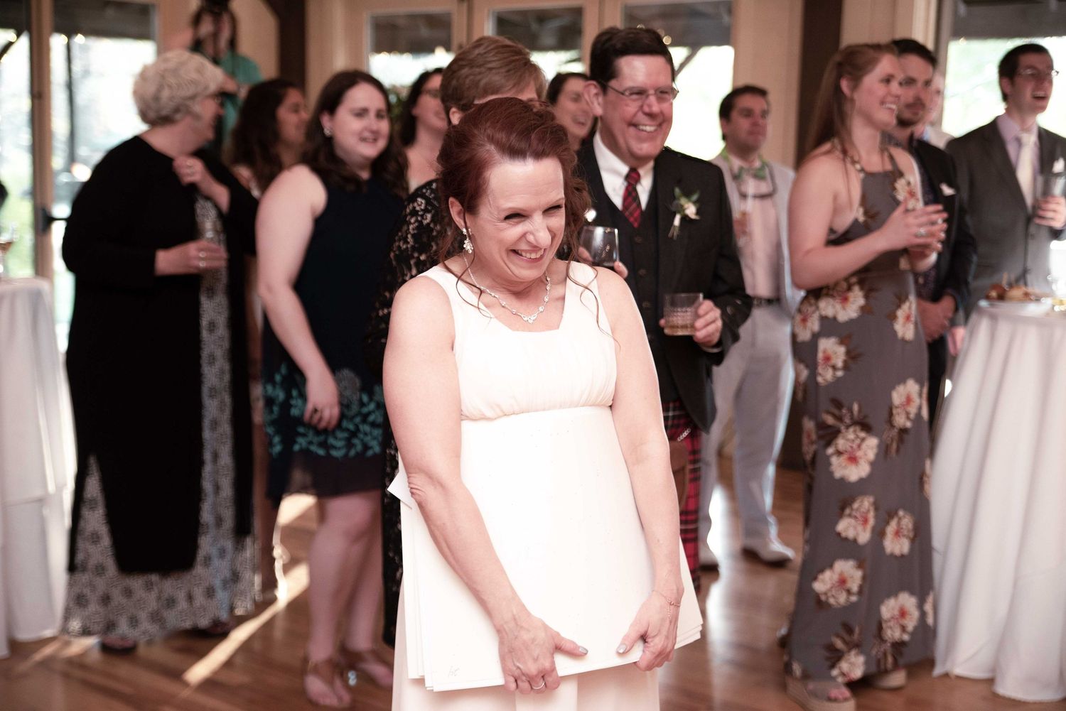 Woman in white dress smiling at a wedding reception with guests gathered in elegant venue.