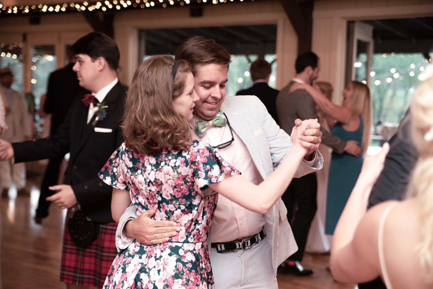 Couple dancing joyfully at a wedding reception, woman in floral dress, man in light suit, guests visible in background.