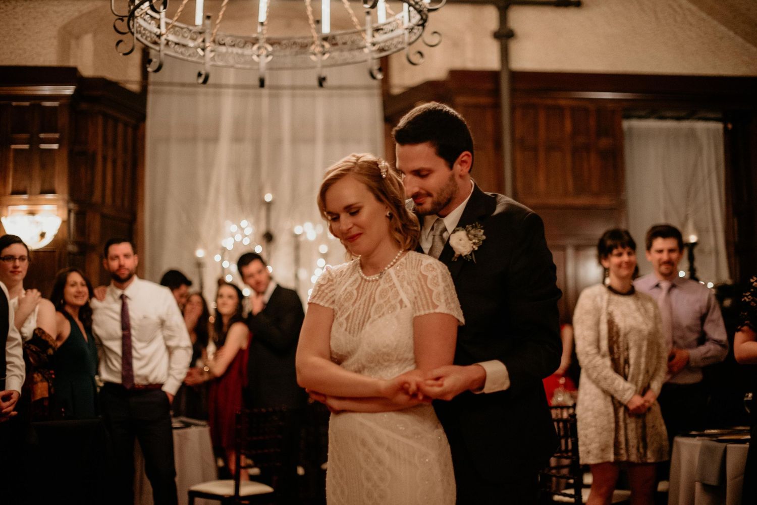 Bride and groom share their first dance surrounded by smiling wedding guests in an elegant chandelier-lit reception hall.