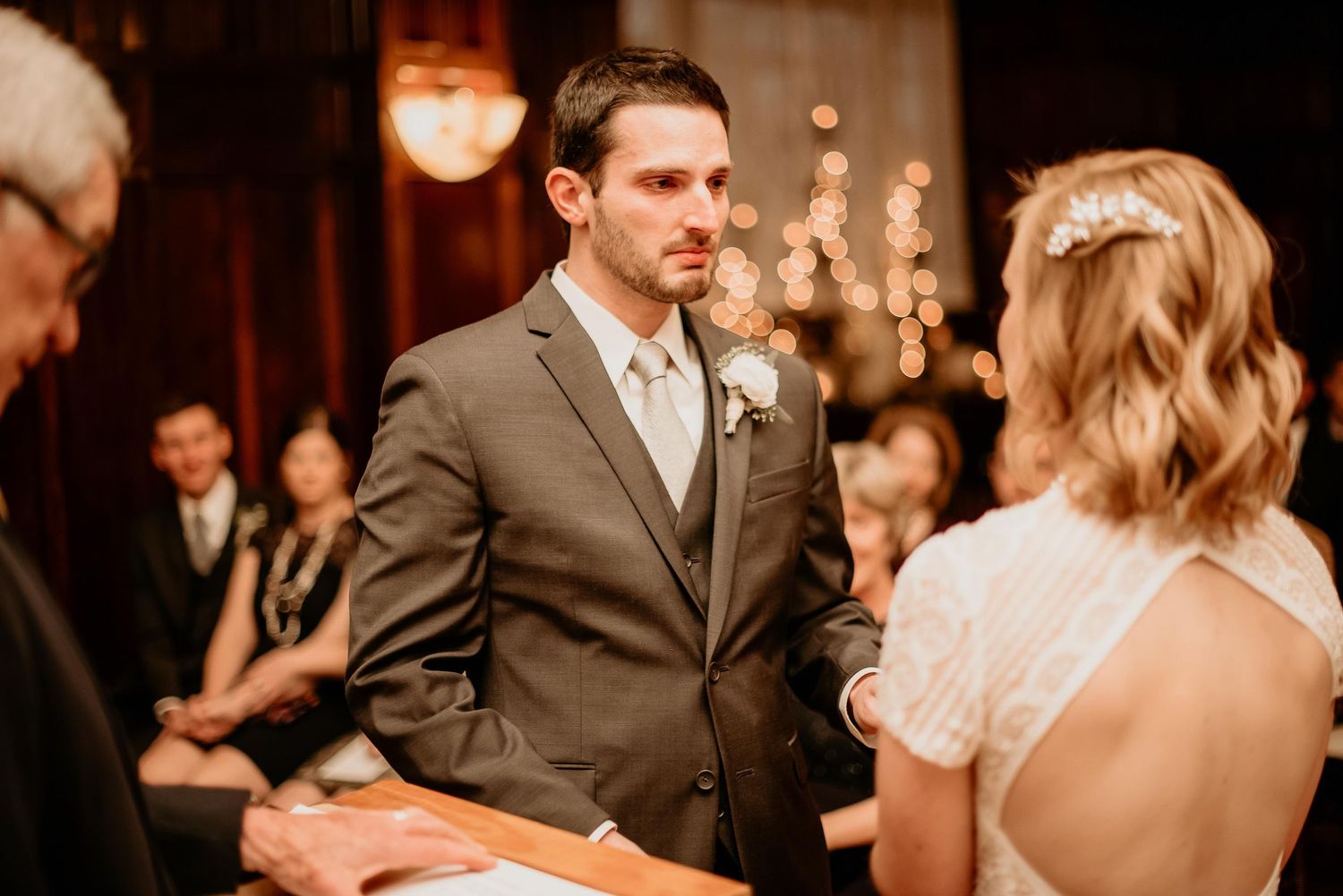 Groom in gray suit exchanges vows with bride in white dress during elegant indoor wedding ceremony.