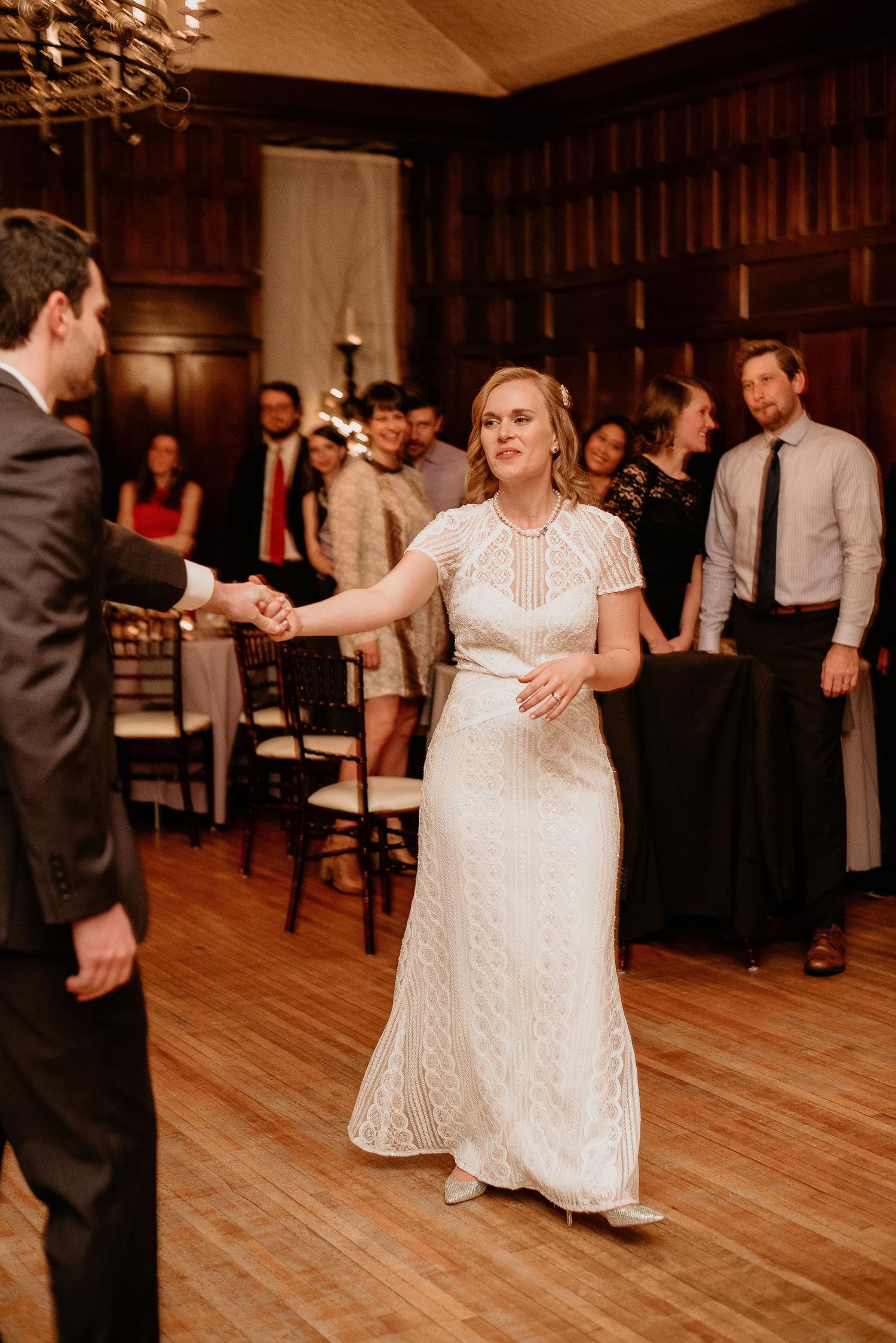 Bride in white lace gown dancing with groom at elegant wood-paneled wedding reception venue.