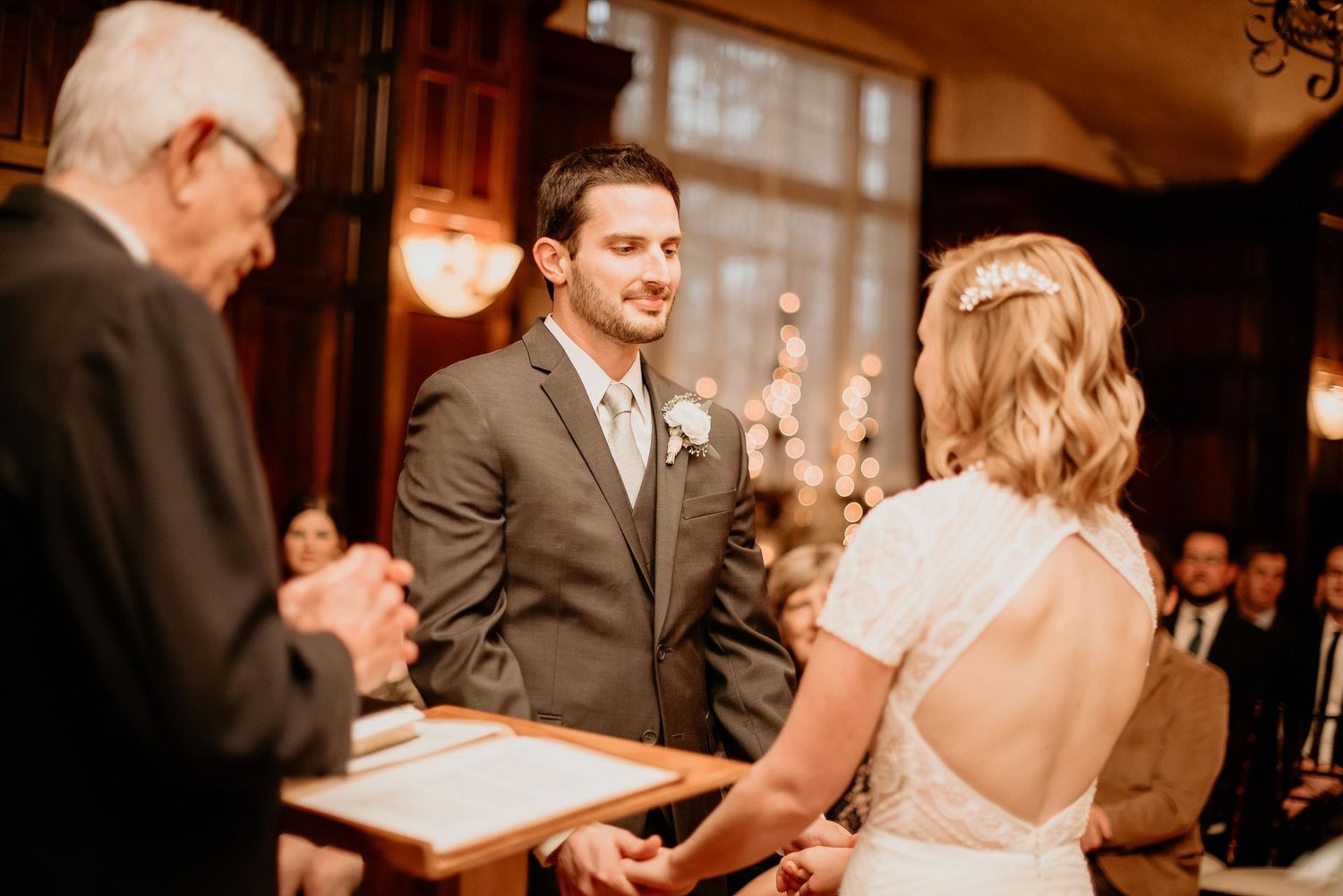 Groom smiling at bride during indoor wedding ceremony while officiant holds book.