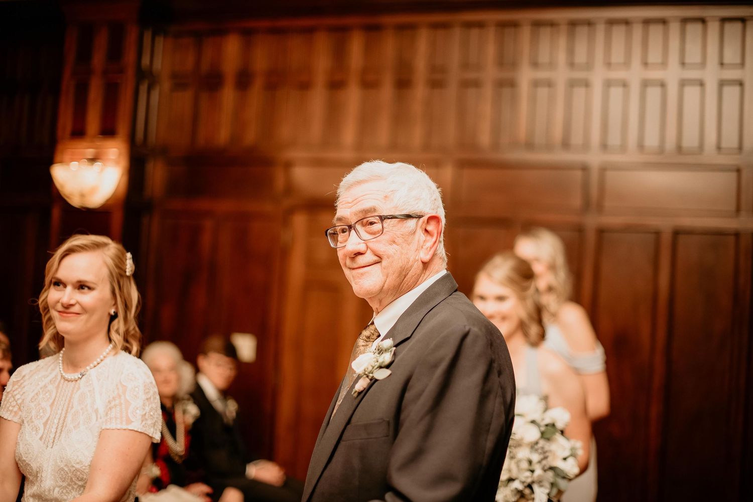 Elderly man smiling warmly at a wedding ceremony, wearing a suit with a boutonniere, standing beside a blonde woman.