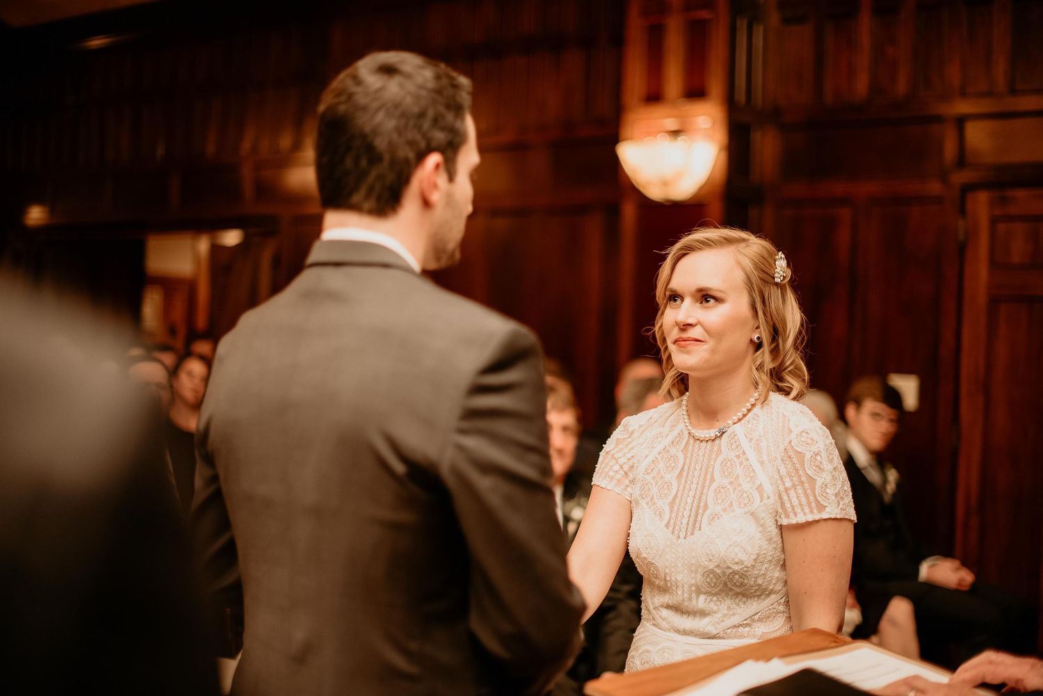 Bride smiling at groom during wedding ceremony in elegant wood-paneled venue with guests in background.