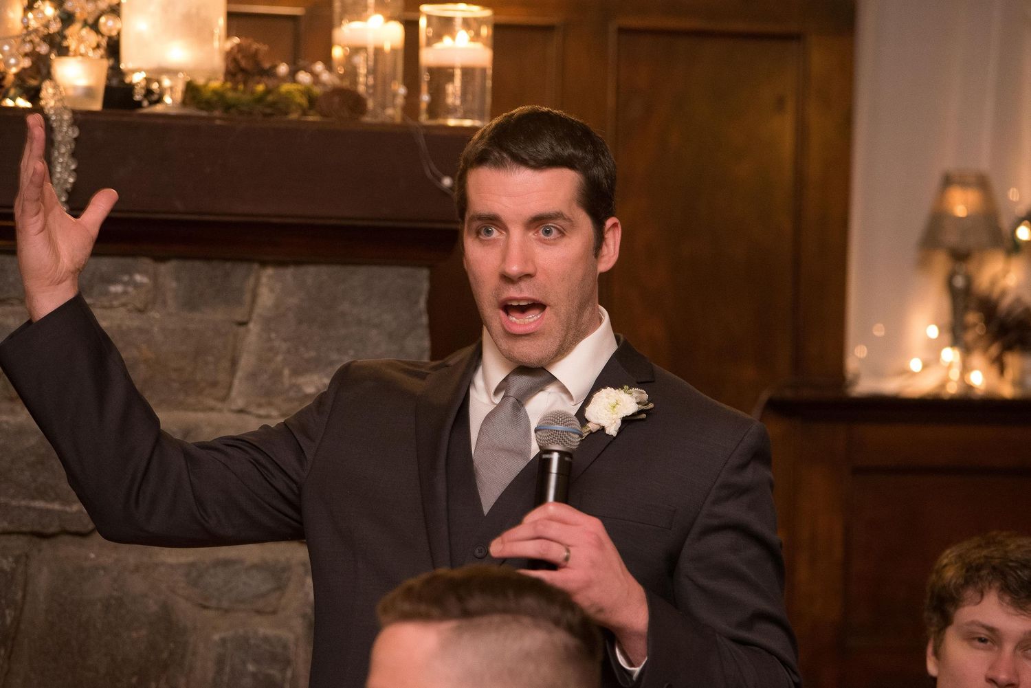 Man in gray suit with boutonniere giving a speech at a wedding reception, holding microphone with hand raised.