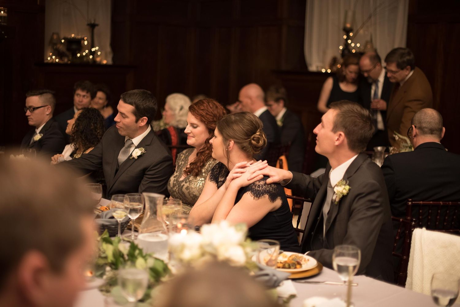 Elegantly dressed wedding guests laughing and socializing at a formal reception dinner table with floral centerpieces.