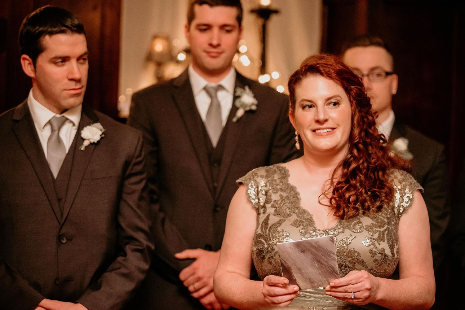 Woman in gold lace dress reads at wedding ceremony with groomsmen in gray suits standing behind her.