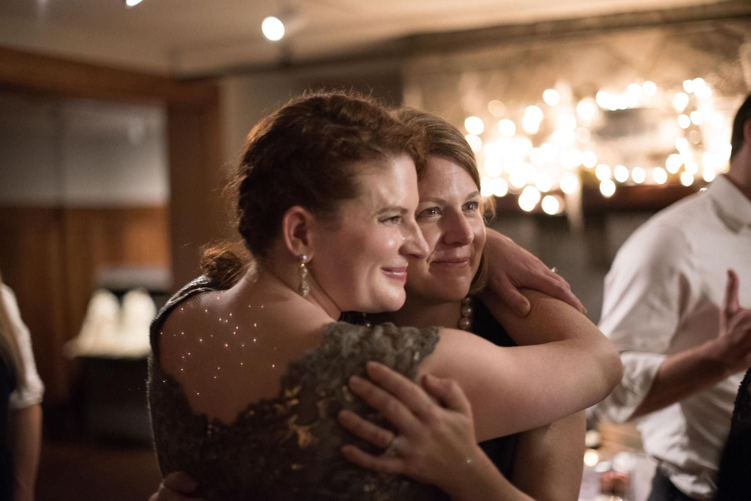Two women share a warm hug at an elegant evening event with soft bokeh lights in the background.