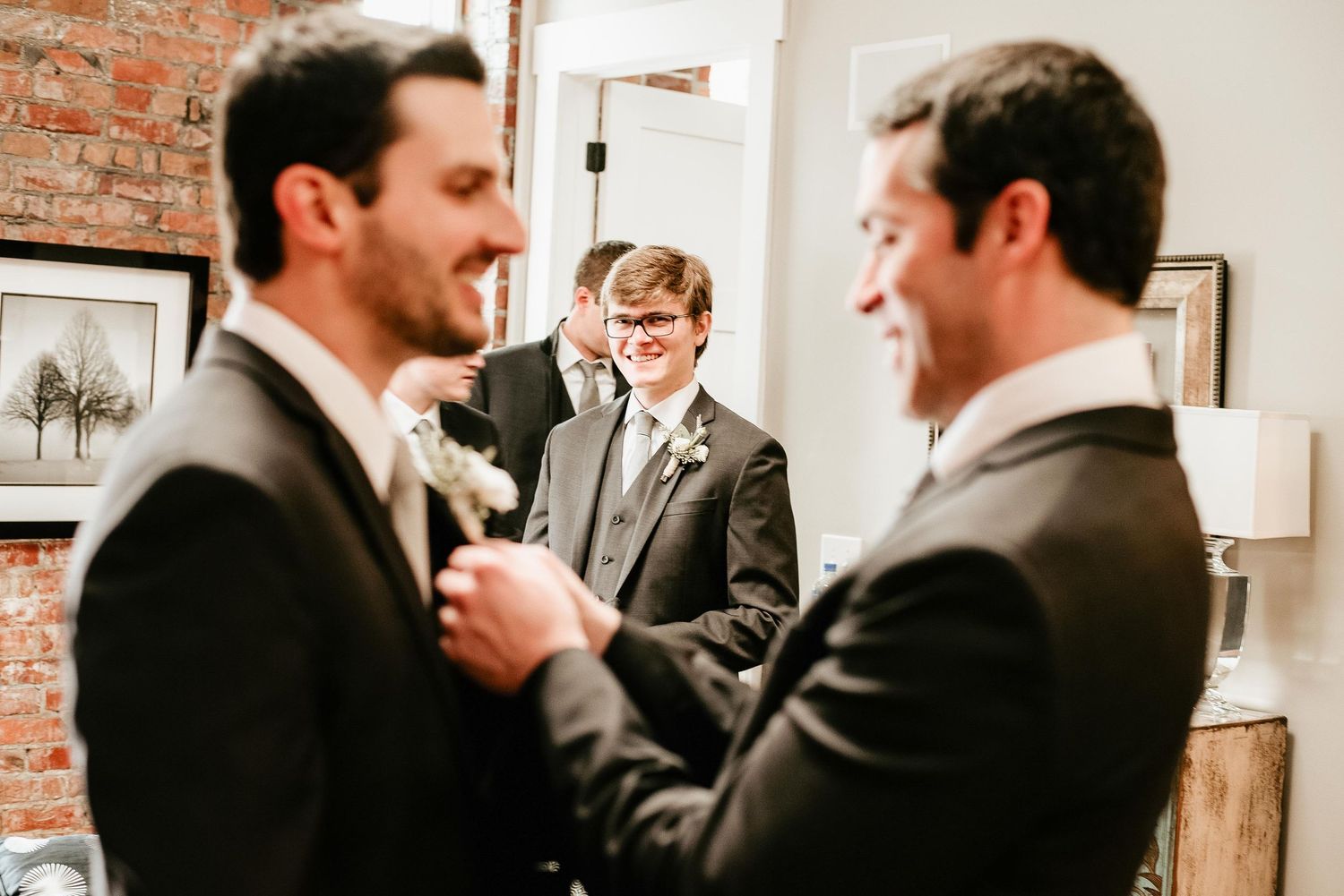 Groomsmen helping groom with boutonniere in rustic brick-walled wedding venue before ceremony.