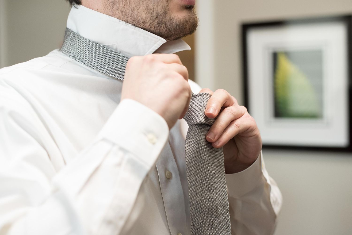 Man in white dress shirt tying a gray necktie, preparing for a formal occasion.